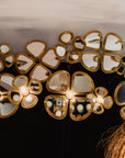 Plusieurs fleurs décoratives en laiton doré à l'effet miroir, fixées au mur et au plafond, certaines équipées d'une ampoule allumée en leur centre, dans une pièce aux murs sombres.