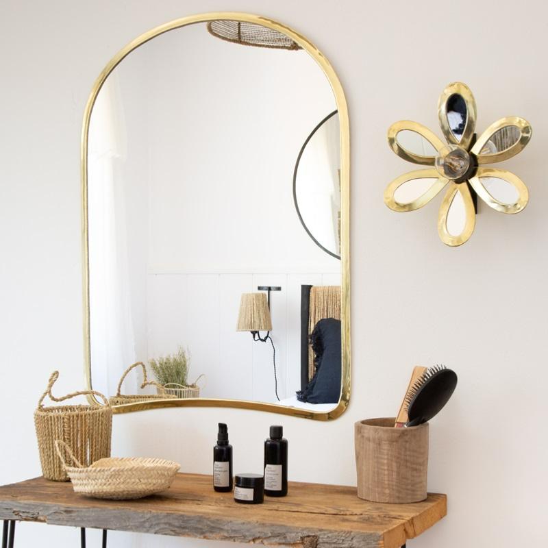 Table en bois brut décorée avec des paniers tressés, des produits de soin et une brosse, surmontée d’un grand miroir doré aux bords arrondis accroché au mur. Une applique murale dorée en forme de fleur est fixée à côté du miroir, créant une ambiance bohème et naturelle.
