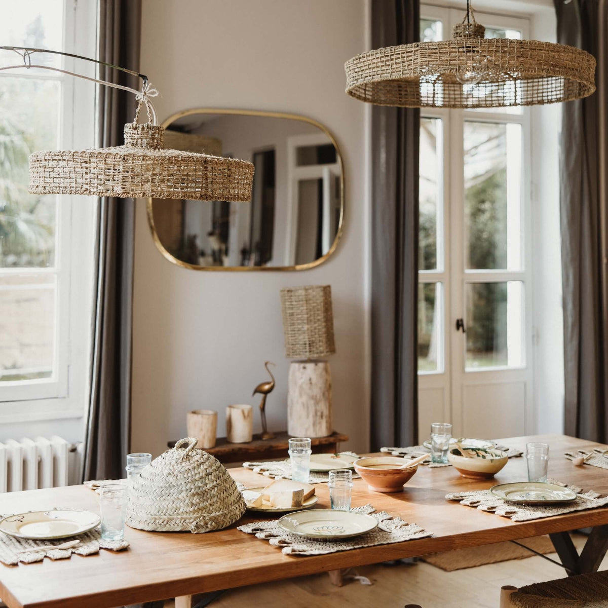Table à manger avec des sets de table et une cloche alimentaire tressés en feuille de palmier, un grand miroir en laiton et des suspensions luminaires tressés en fibre naturelle