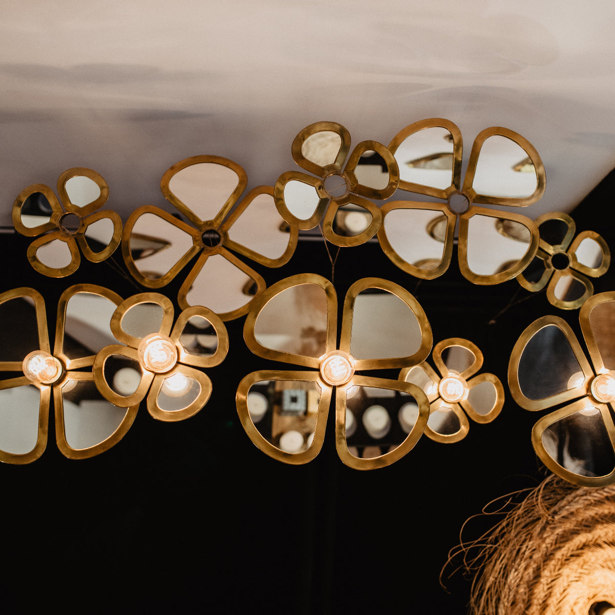 Plusieurs fleurs décoratives en laiton doré à l'effet miroir, fixées au mur et au plafond, certaines équipées d'une ampoule allumée en leur centre, dans une pièce aux murs sombres.