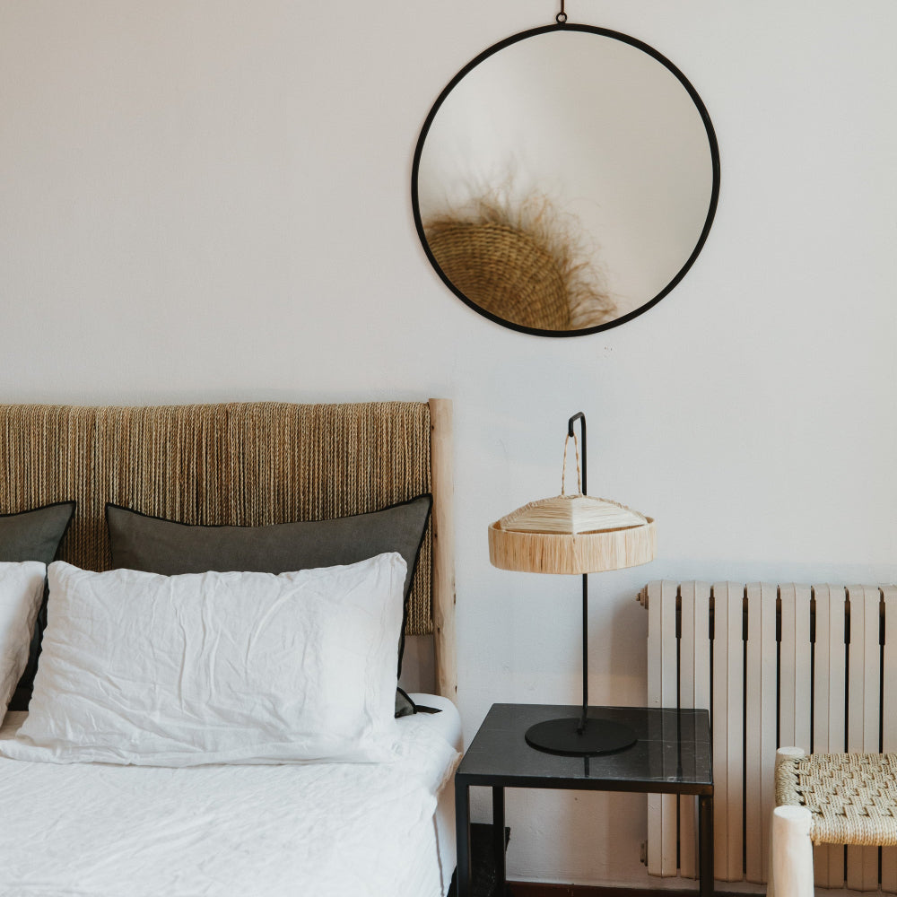 Support de lampe en métal noir avec suspension en fibre naturelle sur une table de chevet, sous un miroir rond