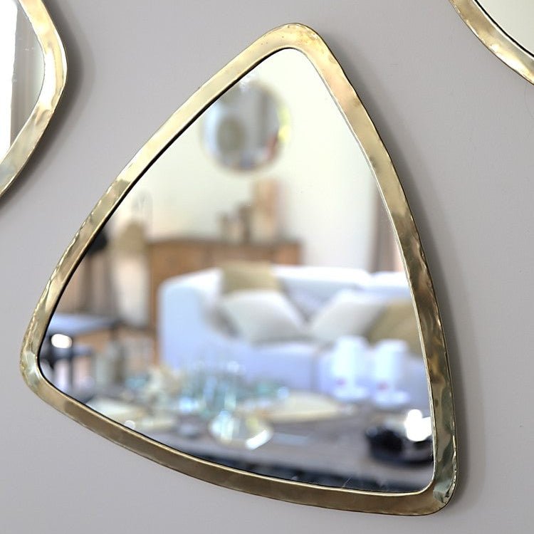 Miroir triangulaire en laiton doré accroché au mur, refletant une pièce lumineuse et naturelle.