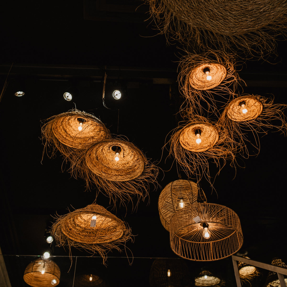 Plusieurs suspensions luminaires en fibre naturelle tressée, allumées et diffusant une lumière dorée, accrochées à différentes hauteurs dans une pièce sombre.